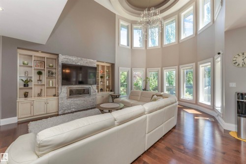 Two-story living area featuring a curved wall of windows, a grand chandelier, hardwood flooring, a stone-clad fireplace, and built-in shelving with cabinetry - 3503 Watson Point(E), Edmonton, AB 