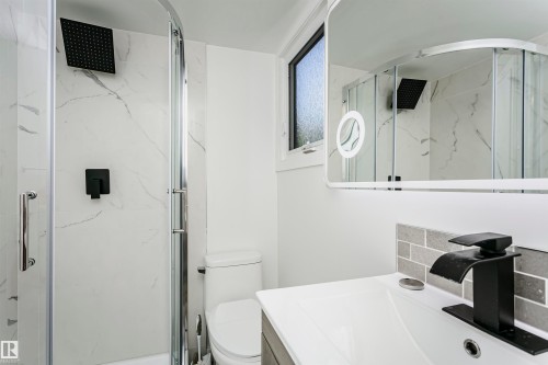 Modern bathroom featuring a glass-enclosed shower with a matte black rain showerhead, a white vanity with an integrated sink, a rectangular illuminated mirror, and a window with privacy glass - 17716 84 Avenue, Edmonton, AB 