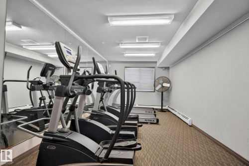 Fitness room featuring multiple cardio machines, full-wall mirror, textured carpet flooring, and fluorescent lighting - 140 301 Clareview Station Drive, Edmonton, AB 
