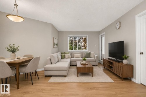 Open-concept living area featuring wood-finish flooring, a contemporary ceiling light fixture, a large window, light-toned walls, and a white door with a silver-tone handle - 140 301 Clareview Station Drive, Edmonton, AB 
