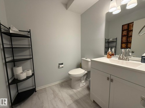 Main Floor 2pc Bathroom featuring light-toned flooring, a white vanity with an integrated sink, a commode, and a large wall-mounted mirror - 166 Roseland Village, Edmonton, AB - Indoor Photo Showing Bathroom
