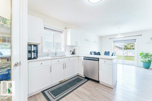 Bright kitchen featuring white cabinetry, stainless steel dishwasher, wood-finish flooring, and a sliding glass door providing access to the backyard - 3525 22 Street, Edmonton, AB - Indoor Photo Showing Kitchen