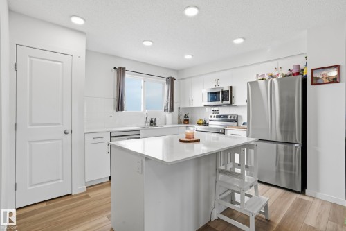 Kitchen featuring wood-finish flooring, white cabinetry, a central island with a white countertop, and stainless steel appliances including a refrigerator, microwave, and range - 58 Jensen Gate, St. Albert, AB 