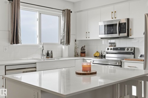 Contemporary kitchen featuring white cabinetry, a large center island with a light-toned countertop, stainless steel appliances, a farmhouse-style sink, and a white tiled backsplash - 58 Jensen Gate, St. Albert, AB 