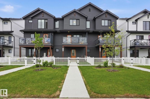 Contemporary multi-level featuring dark-toned vertical and horizontal siding, a private balcony with glass railing, and a white picket fence enclosing a landscaped front yard with a concrete walkway - 58 Jensen Gate, St. Albert, AB 