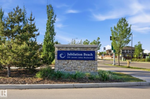 Stone entry monument with a blue placque featuring "Jubilation Beach" and "205 Jensen Lakes Boulevard." The monument is surrounded by manicured landscaping including evergreen trees and ornamental grasses - 58 Jensen Gate, St. Albert, AB 