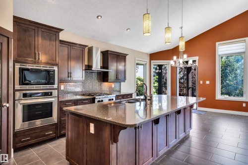 Modern kitchen featuring dark wood cabinetry, stainless steel appliances, a center island with sink and stone countertop, tile flooring, and high vaulted ceilings - 9 Shorewood Crescent, Leduc, AB - Indoor Photo Showing Kitchen With Upgraded Kitchen