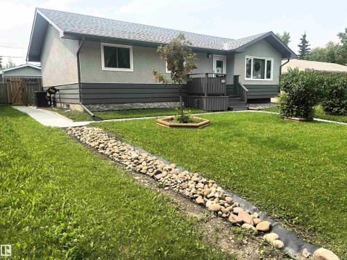 Stucco exterior with horizontal dark grey siding accents, a prominent bay window, a front porch with dark grey railing and steps, and a landscaped front yard featuring a rock-lined channel - 4812 47 St, Drayton Valley, AB 