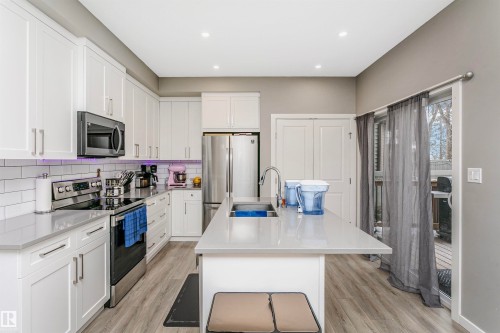 Modern kitchen featuring white cabinetry, stainless steel appliances, a large center island with an undermount sink, light gray countertops, and wood-finish flooring - 7322 Chivers Crescent, Edmonton, AB 