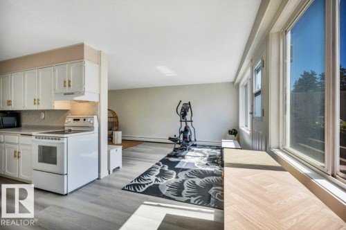 Open-concept living area featuring light wood-finish flooring, a kitchen with white cabinetry and gold-tone hardware, a white electric range, and large windows with a herringbone-patterned windowsill - 102 7805 159 Street, Edmonton, AB 