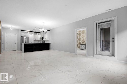 Open-concept living area featuring large format tile flooring and a kitchen with white cabinetry, stainless steel appliances, and a dark island - 105 160 Magrath Road, Edmonton, AB 