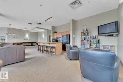 Spacious common area featuring textured carpeting, a kitchen area with light wood cabinetry, and a peninsula with bar seating - 105 160 Magrath Road, Edmonton, AB 