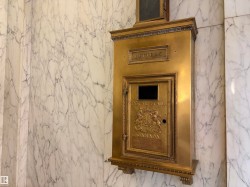 Brass mail drop box with embossed details, set into a wall of white marble with gray veining - 