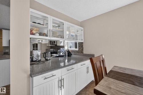 Kitchen featuring white shaker-style cabinetry with brushed nickel hardware, glass-front upper cabinets, speckled gray countertops, and a mosaic tile backsplash - 2327 37B Avenue, Edmonton, AB 