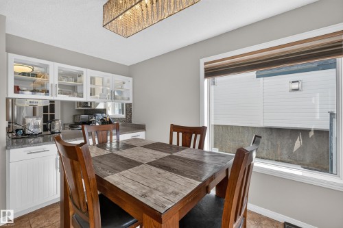 Dining area featuring a modern rectangular chandelier, built-in cabinetry with glass-front upper cabinets, a granite-finish countertop, and a large window with a roller shade - 2327 37B Avenue, Edmonton, AB 