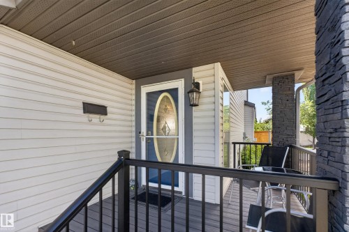 Covered porch featuring light-colored siding, a dark blue entry door with an oval glass insert, a dark gray wood-finish deck, black metal railings, and a stone-clad support pillar - 2327 37B Avenue, Edmonton, AB 