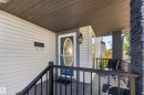 Covered porch featuring light-colored siding, a dark blue entry door with an oval glass insert, a dark gray wood-finish deck, black metal railings, and a stone-clad support pillar - 2327 37B Avenue, Edmonton, AB 