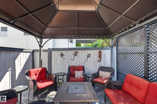 Covered outdoor patio area featuring a dark-framed gazebo structure with overhead lighting, a built-in fire pit table, light-toned wood fencing, and dark lattice privacy screens - 2327 37B Avenue, Edmonton, AB 