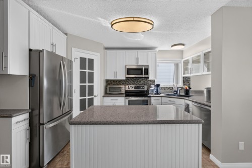 Kitchen featuring a central island with beadboard paneling, light-toned countertops, white cabinetry, stainless steel appliances, and a mosaic tile backsplash - 2327 37B Avenue, Edmonton, AB 