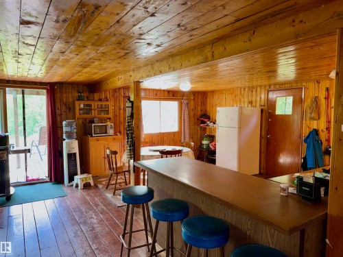 Open-concept living space with wood-paneled walls and ceiling, featuring a kitchen area with a built-in cabinet, a breakfast bar, and a sliding glass door opening to an exterior deck - 208 60118 Secondary Highway 867, Rural St. Paul County, AB - Indoor