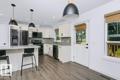 Bright kitchen featuring wood-finish flooring, white cabinetry, a light-toned countertop island with pendant lighting, and a full suite of black appliances - 27 4205 30 Street, Edmonton, AB 