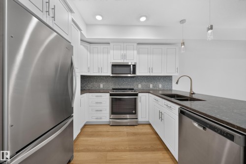 Kitchen featuring wood-finish flooring, white cabinetry with brushed nickel hardware, stainless steel appliances, dark countertops, and a mosaic tile backsplash - Sherwood Park, AB 