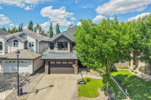 Modern residential exterior featuring a stone and stucco facade, a dark shingle roof, a two-car garage, and a mature front yard tree - 1231 Tredger Crest, Edmonton, AB 