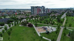 Expansive green space featuring a playground with multiple structures, concrete pathways, mature trees, and surrounding residential properties including multi-story buildings and single-family homes - 