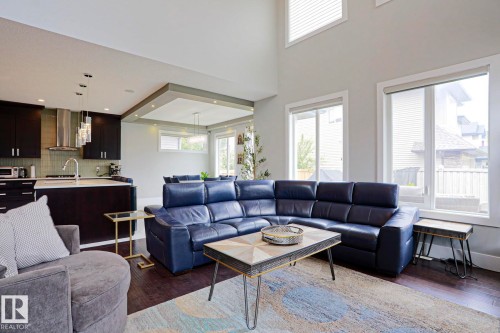Open-concept living space featuring wood-finish flooring, a two-story ceiling, multiple windows, a kitchen island with seating, and dark cabinetry - 1304 Ainslie Wynd, Edmonton, AB 