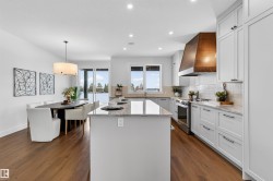 Open-concept kitchen and dining area featuring wood-finish flooring, a central island with a light-toned countertop, white cabinetry with dark hardware, and a wood-paneled range hood - 