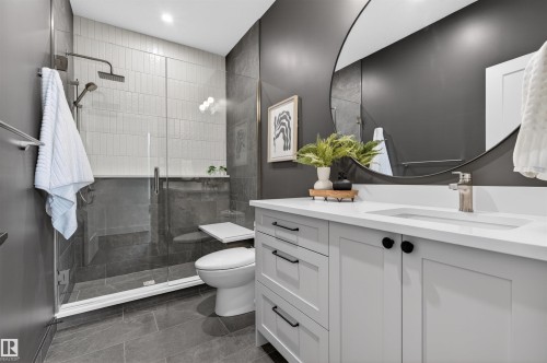 Modern bathroom featuring a frameless glass shower enclosure with a rain showerhead, a white rectangular sink with a single-handle faucet, light gray vanity cabinetry with black hardware, a round frameless mirror, and dark gray floor tiling - 149 Eastgate Way, St. Albert, AB 