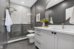 Modern bathroom featuring a frameless glass shower enclosure with a rain showerhead, a white rectangular sink with a single-handle faucet, light gray vanity cabinetry with black hardware, a round frameless mirror, and dark gray floor tiling - 