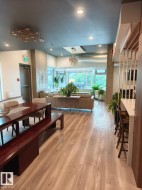 Open-concept area featuring light wood-finish flooring, a dark wood dining table with bench seating, a contemporary chandelier, and large windows providing natural light - 