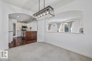 Carpeted dining area with a linear chandelier, arched passthrough, and a half wall opening to a kitchen with wood-finish flooring, a kitchen island, and white cabinetry - 2407 Hagen Way, Edmonton, AB 