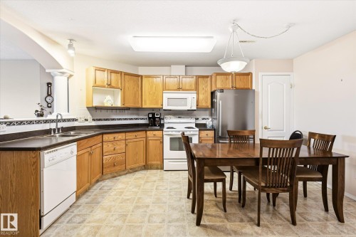 Open-concept kitchen featuring wood cabinetry, white appliances, and a stainless steel refrigerator - #310 237 Youville Dr E Nw, Edmonton, AB - Indoor Photo Showing Kitchen With Double Sink