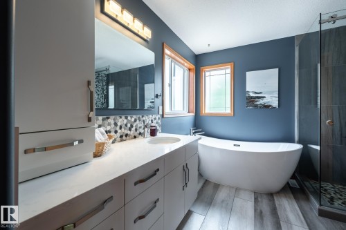 Contemporary ensuite bathroom featuring a freestanding soaking tub, glass-enclosed shower, wood-finish tile flooring, a dual-sink vanity with a light-colored countertop, and a mosaic tile backsplash - 114 Reichert Drive, Beaumont, AB 