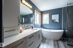 Contemporary ensuite bathroom featuring a freestanding soaking tub, glass-enclosed shower, wood-finish tile flooring, a dual-sink vanity with a light-colored countertop, and a mosaic tile backsplash - 