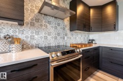 Contemporary kitchen featuring dark wood-finish cabinetry, light-toned countertops, a patterned tile backsplash, stainless steel range with overhead hood, and under-cabinet lighting - 