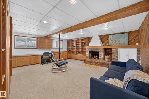 Spacious lower-level room featuring a brick fireplace with a wood mantel, extensive built-in shelving, and custom cabinetry with integrated desk space - 3411 103 Avenue, Edmonton, AB 