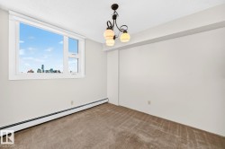Spacious room featuring a large window with city skyline, neutral carpeting, white painted walls, baseboard heating, and an ornate ceiling light fixture - 