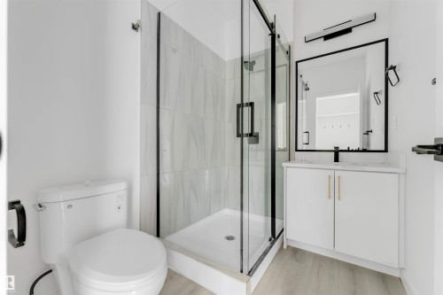 Bathroom featuring a glass-enclosed shower with tiled walls, a white vanity with gold-tone hardware, a rectangular framed mirror, and light wood-finish flooring - 3605 112 Ave, Edmonton, AB 