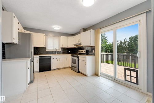 Kitchen featuring white cabinetry, dark tile backsplash, stainless steel refrigerator, black dishwasher, and a sliding glass door opening to an outdoor deck - 193 Woodvale Road W, Edmonton, AB 