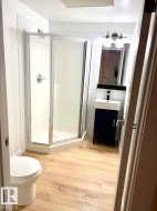 Compact bathroom featuring wood-finish flooring, a corner shower enclosure, white subway tile shower surround, a contemporary vanity with an integrated sink, and a wall-mounted light fixture above the vanity mirror - 