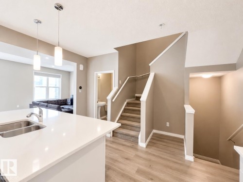 Open-concept interior featuring light wood-finish flooring, a contemporary staircase with carpeted treads, a kitchen island with a double stainless steel sink, and modern pendant lighting - 20104 19 Avenue, Edmonton, AB 
