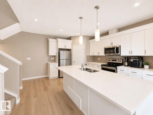 Bright kitchen featuring a large central island with dual pendant lighting, white cabinetry, integrated stainless steel appliances, a mosaic tile backsplash, and wood-finish flooring - 20104 19 Avenue, Edmonton, AB 