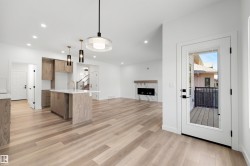 Open-concept main living area featuring wood-finish flooring throughout, a modern kitchen island with a quartz countertop, recessed lighting, a contemporary fireplace with a stone surround, and a glass-paneled door leading to an outdoor deck - 