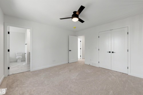 Spacious carpeted room featuring a ceiling fan with integrated lighting, dual white panel doors with matte black hardware, and an en-suite bathroom with marble-look tile flooring - 30 Elsinore Place, Edmonton, AB 