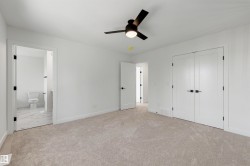 Spacious carpeted room featuring a ceiling fan with integrated lighting, dual white panel doors with matte black hardware, and an en-suite bathroom with marble-look tile flooring - 