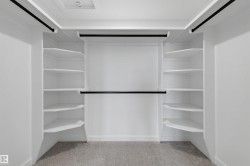Spacious walk-in closet featuring white built-in shelving, multiple hanging rods, and light gray carpet flooring - 