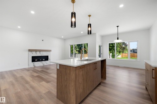 Open-concept living area featuring a kitchen island with a gold-tone faucet, wood-finish flooring, a modern fireplace with a marble-patterned surround, recessed lighting, and large windows - 30 Elsinore Place, Edmonton, AB 